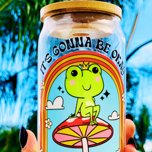 Load image into Gallery viewer, A 16 oz glass cup featuring a colorful UVDTF wrap with a smiling frog on a mushroom under a rainbow and the text “It’s Gonna Be Okay.” Comes with a bamboo lid and reusable glass straw. BPA-free and handwash only.