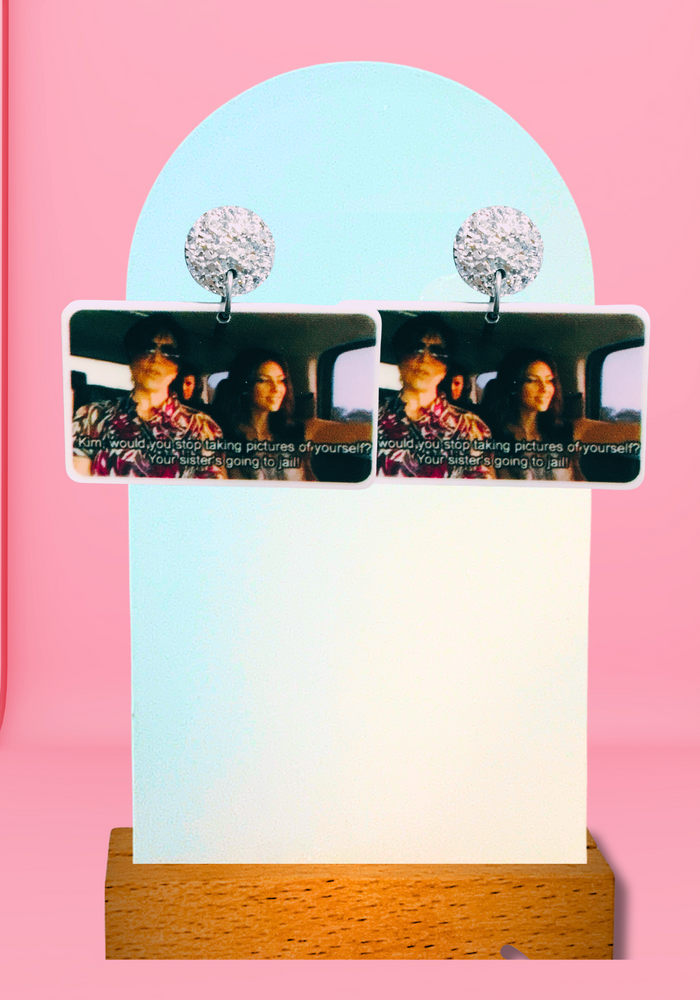 Pink backdrop with earrings featuring the memorable scene from Keeping Up with the Kardashians where Kim is told to stop taking photos while her sister is going to jail.