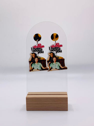 Handcrafted acrylic earrings showcasing the iconic '10 Things I Hate About You' movie poster design. The earrings feature a vibrant image featuring Julia Stiles leaning against Heath Ledger. The bold typography says 