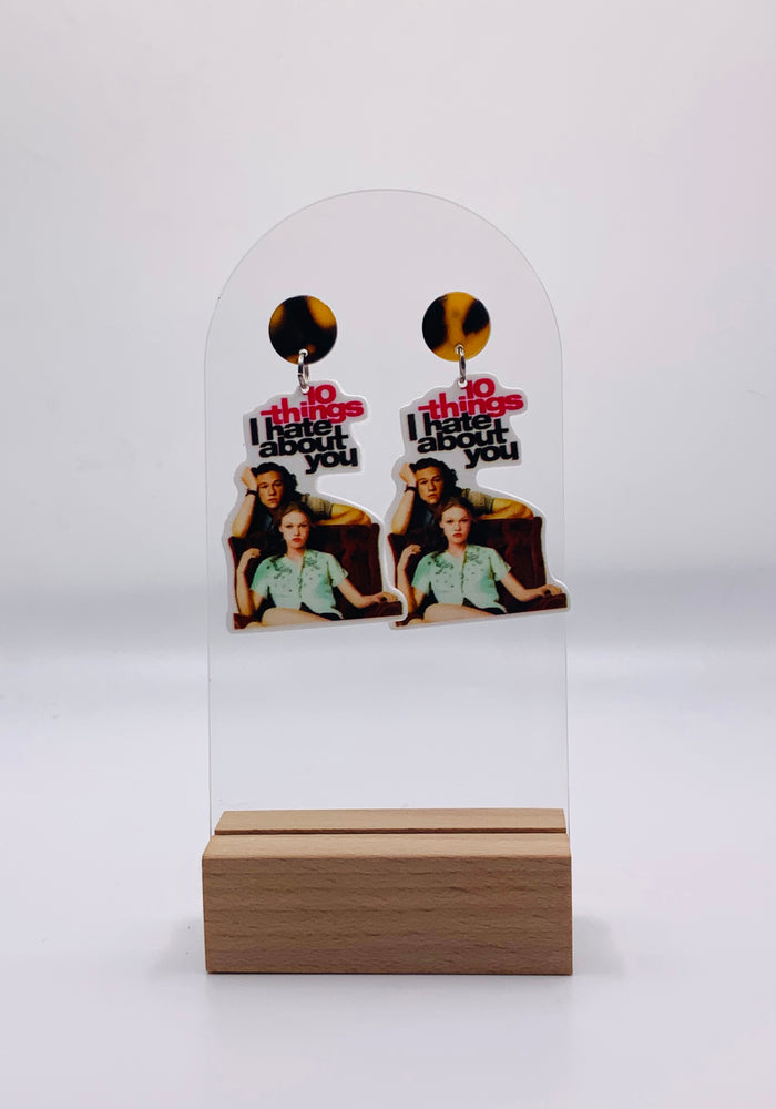 Handcrafted acrylic earrings showcasing the iconic '10 Things I Hate About You' movie poster design. The earrings feature a vibrant image featuring Julia Stiles leaning against Heath Ledger. The bold typography says 