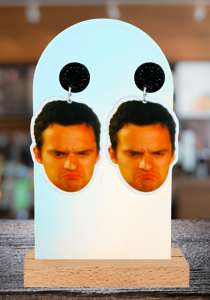 A bar background with a pair of New Girl earrings featuring Nick Miller’s grumpy frowning face