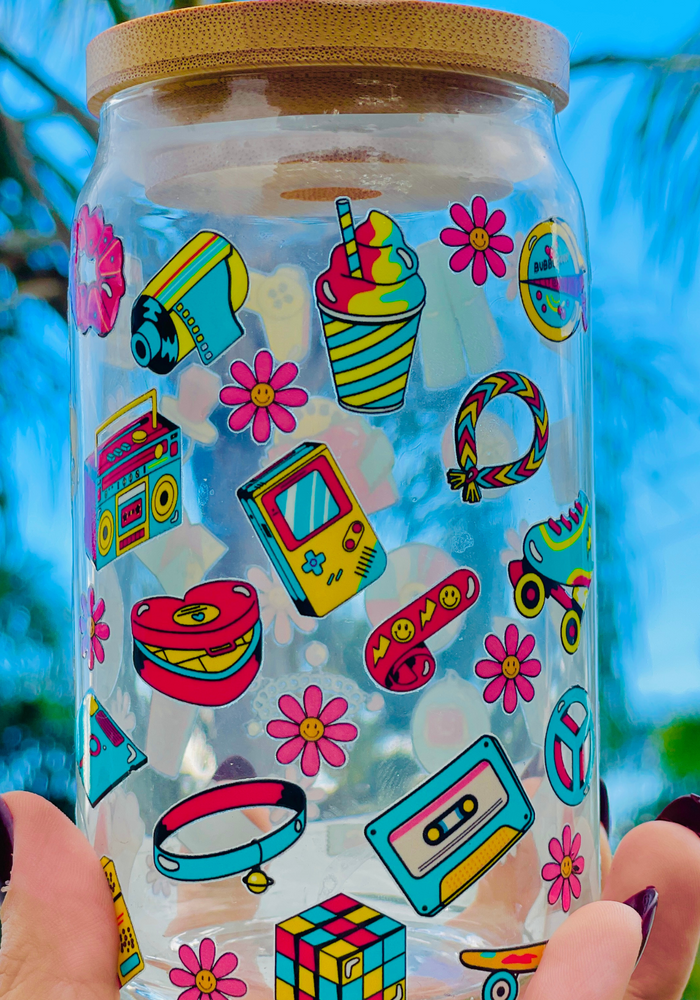 A 16oz BPA-free glass cup featuring a colorful 90s-themed UVDTF wrap with icons like Game Boys, cassette tapes, roller skates, and boomboxes. Comes with a bamboo lid and reusable glass straw, perfect for nostalgic drinkware lovers.