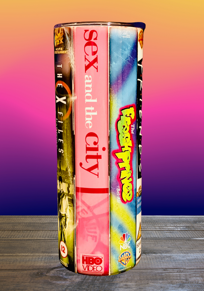 A 20 oz stainless steel insulated tumbler featuring a vinyl wrap of VHS covers from iconic '90s TV shows, including Friends, The Sopranos, The Fresh Prince of Bel-Air, and Sex and the City. Comes with a lid and reusable straw, perfect for nostalgic TV lovers.