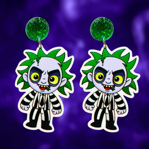 A purple background with earrings featuring a cute cartoon version of Beetlejuice, perfect for fans of the classic film and nostalgic Halloween fun.