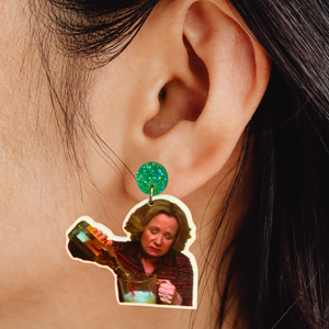 A close-up of an ear wearing a green gem stud with an earring of Kitty Forman from That 70s Show pouring a bottle of liquor into a blender