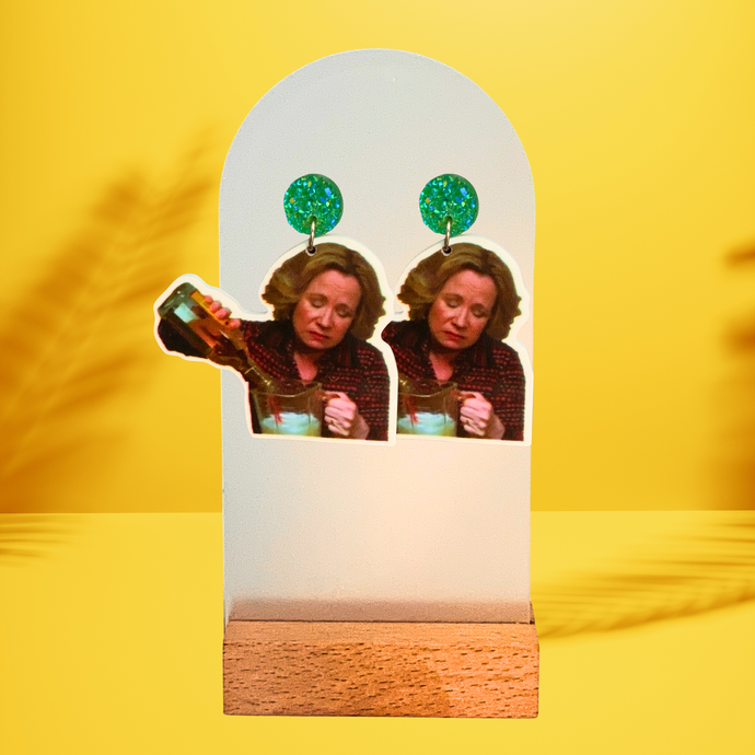 Vibrant yellow background featuring a wooden earring stand with images on green gems featuring Kitty Foreman pouring an entire bottle of liquor into a blender