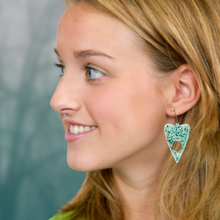 Load image into Gallery viewer, A blonde model wearing a pair of aqua blue translucent Ouija planchette earrings, perfect for adding a fun and mystical touch to your Halloween look.
