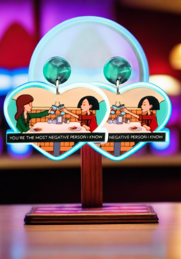 Earrings sitting on a stand that feature Daria and Jane cheersing with the caption 