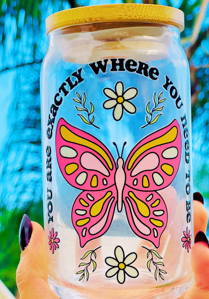 A 16 oz glass cup featuring a UVDTF wrap with a pink butterfly, daisies, and the phrase “You’re Exactly Where You Need To Be.” Comes with a bamboo lid and reusable glass straw. BPA-free and handwash only.