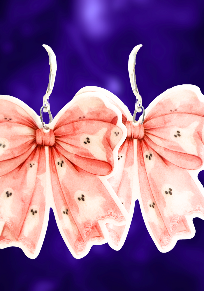 A purple background with pink coquette bow earrings with adorable ghost patterns, perfect for adding a cute and spooky twist to Halloween fashion.