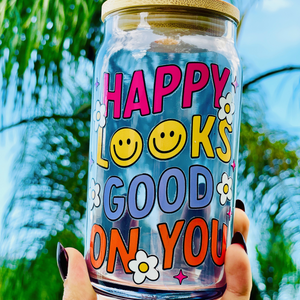 A 16oz BPA-free glass cup featuring a retro-inspired UVDTF design with smiley faces, daisies, and the phrase "Happy Looks Good on You," complete with a bamboo lid and reusable glass straw.