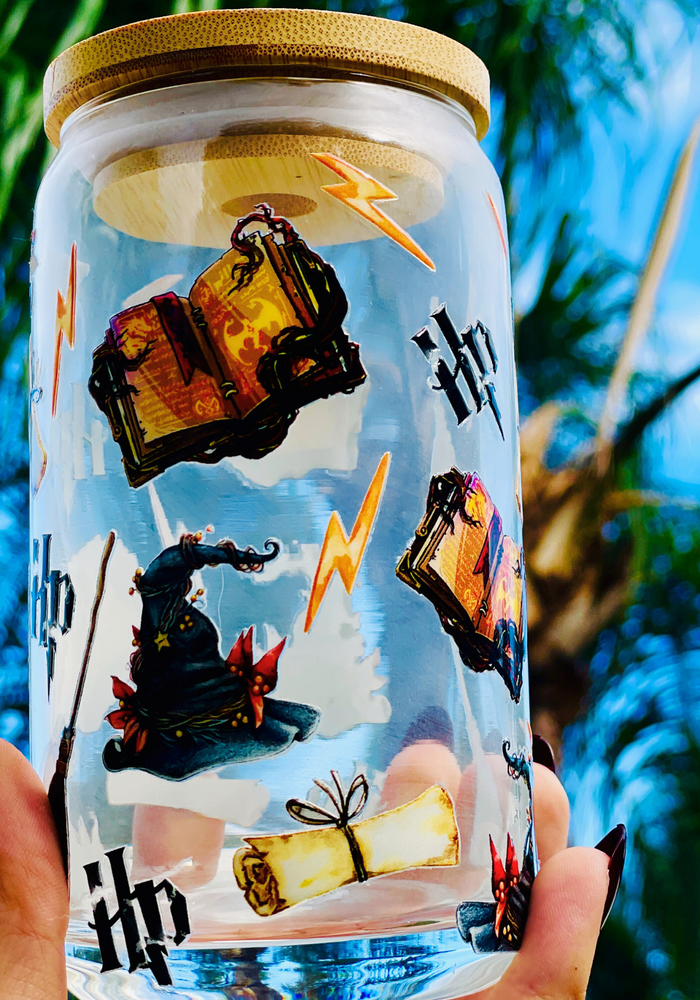 A 16 oz glass cup featuring a Harry Potter-inspired UVDTF wrap with wands, spellbooks, lightning bolts, and the Sorting Hat. Includes a bamboo lid and reusable glass straw, perfect for cold drinks. BPA-free and handwash only.