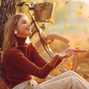 A woman sitting in front of a tree with falling autumn leaves, wearing earrings with the phrase 'Hello Autumn' featuring doodles of a scarf, sweater, pumpkin, leaves, coffee, candles, and pumpkin pie, perfect for cozy fall enthusiasts 