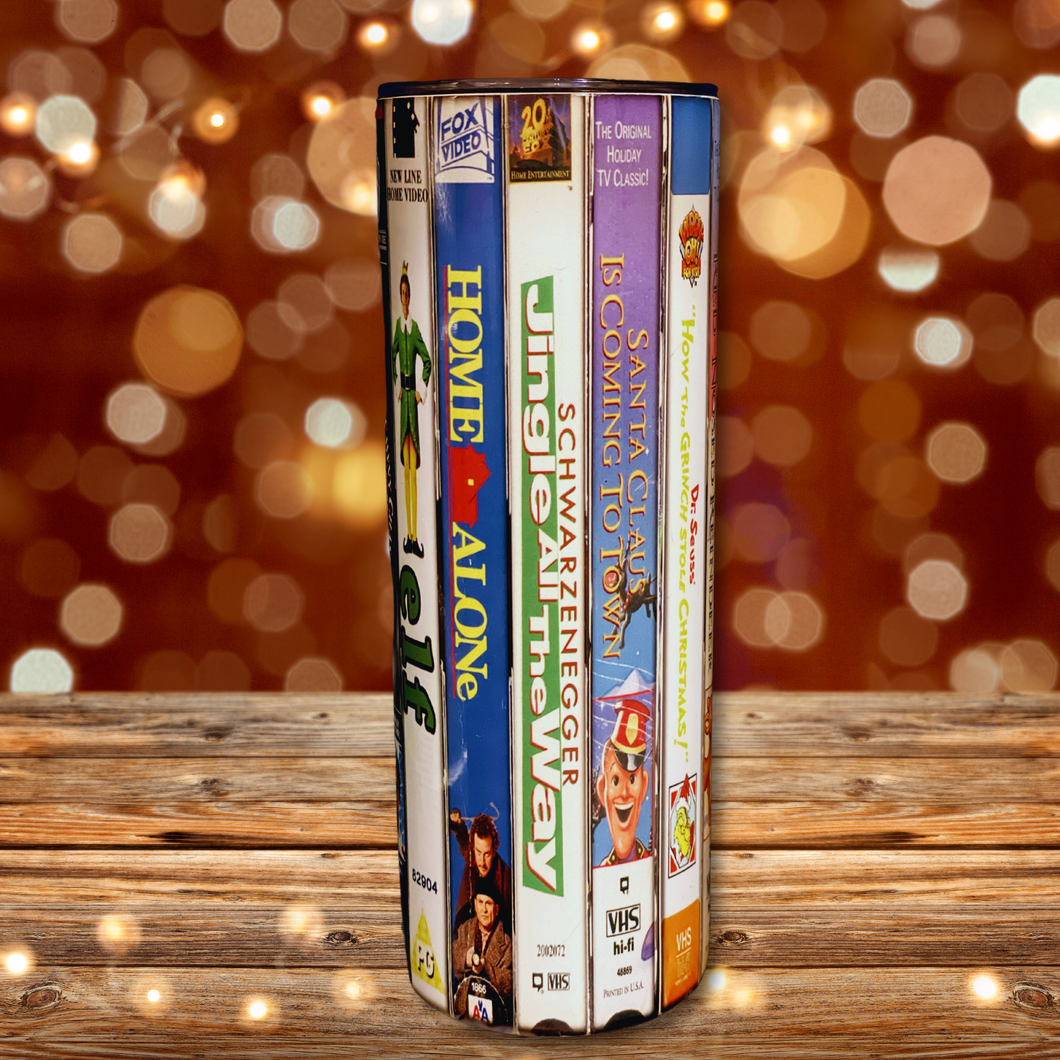 A 20 oz stainless steel insulated Christmas Movie Tumbler featuring a colorful design with movie covers including Elf, Home Alone, Jingle All The Way, and Santa Claus is Coming to Town. Comes with a clear lid, metal straw, and straw cleaner, perfect for hot and cold drinks.