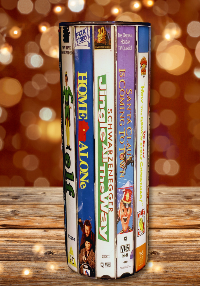 A 20 oz stainless steel insulated Christmas Movie Tumbler featuring a colorful design with movie covers including Elf, Home Alone, Jingle All The Way, and Santa Claus is Coming to Town. Comes with a clear lid, metal straw, and straw cleaner, perfect for hot and cold drinks.