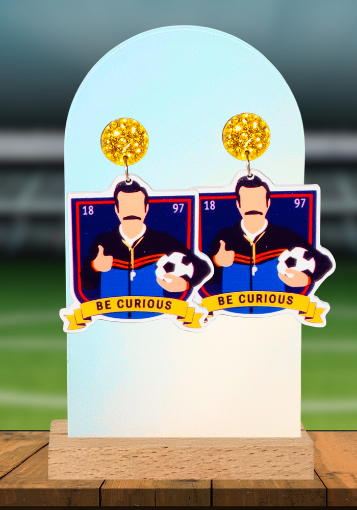 Ted Lasso “be curious” earrings displayed on a tabletop on a soccer field