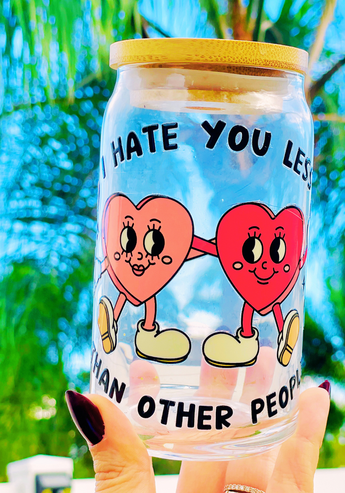A 16 oz glass cup featuring a UVDTF wrap with two hand-holding hearts and the phrase “I Hate You Less Than Other People.” Comes with a bamboo lid and reusable glass straw, perfect for cold drinks. BPA-free and handwash only.