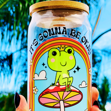 Load image into Gallery viewer, A 16 oz glass cup featuring a colorful UVDTF wrap with a smiling frog on a mushroom under a rainbow and the text “It’s Gonna Be Okay.” Comes with a bamboo lid and reusable glass straw. BPA-free and handwash only.
