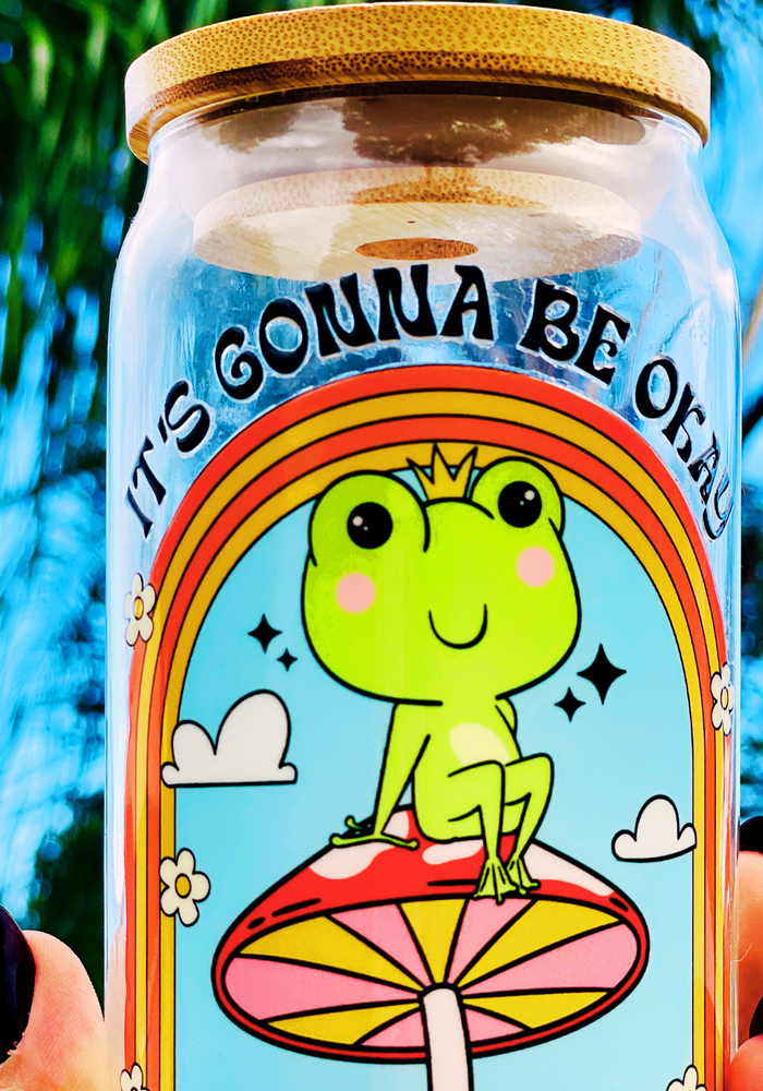 A 16 oz glass cup featuring a colorful UVDTF wrap with a smiling frog on a mushroom under a rainbow and the text “It’s Gonna Be Okay.” Comes with a bamboo lid and reusable glass straw. BPA-free and handwash only.