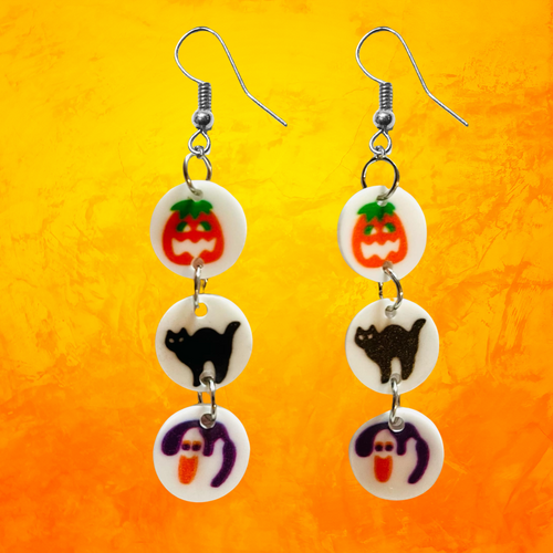 Earrings featuring mini replicas of Pillsbury Halloween sugar cookies, including jack-o'-lanterns, black cats, and ghosts, dangling from hooks against an orange background. Perfect for Halloween nostalgia and fun fall fashion.