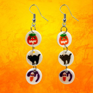 Earrings featuring mini replicas of Pillsbury Halloween sugar cookies, including jack-o'-lanterns, black cats, and ghosts, dangling from hooks against an orange background. Perfect for Halloween nostalgia and fun fall fashion.