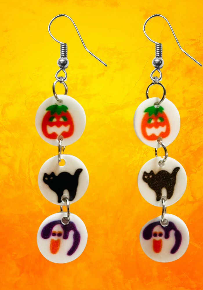 Earrings featuring mini replicas of Pillsbury Halloween sugar cookies, including jack-o'-lanterns, black cats, and ghosts, dangling from hooks against an orange background. Perfect for Halloween nostalgia and fun fall fashion.
