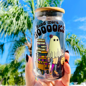 A 16 oz glass cup featuring a cute ghost design pulling a wagon full of books with the text "Read More BOOks." Includes a bamboo lid decorated with tiny ghosts and a reusable glass straw, ideal for book lovers.