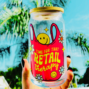 A vibrant 20 oz BPA-free glass cup with a bold design reading “Time for that Retail Therapy,” featuring a pink tote bag, smiley face, daisies, and stars. Comes with a bamboo lid and reusable glass straw.