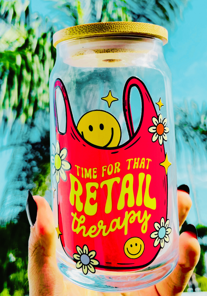 A vibrant 20 oz BPA-free glass cup with a bold design reading “Time for that Retail Therapy,” featuring a pink tote bag, smiley face, daisies, and stars. Comes with a bamboo lid and reusable glass straw.