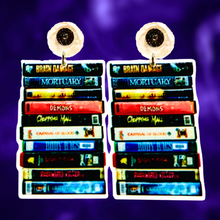 Load image into Gallery viewer, A purple background with earrings featuring a stack of classic cult horror VHS tapes, including Brain Damage, Firestarter, and Chopping Mall, ideal for fans of vintage horror movies.
