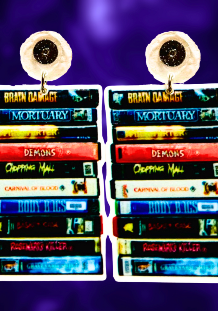 A purple background with earrings featuring a stack of classic cult horror VHS tapes, including Brain Damage, Firestarter, and Chopping Mall, ideal for fans of vintage horror movies.